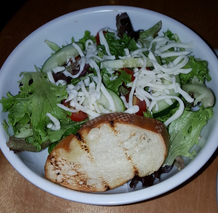 A house salad at Sagra features shredded cheese and a piece of toasted bread. It is served with a choice of dressing.