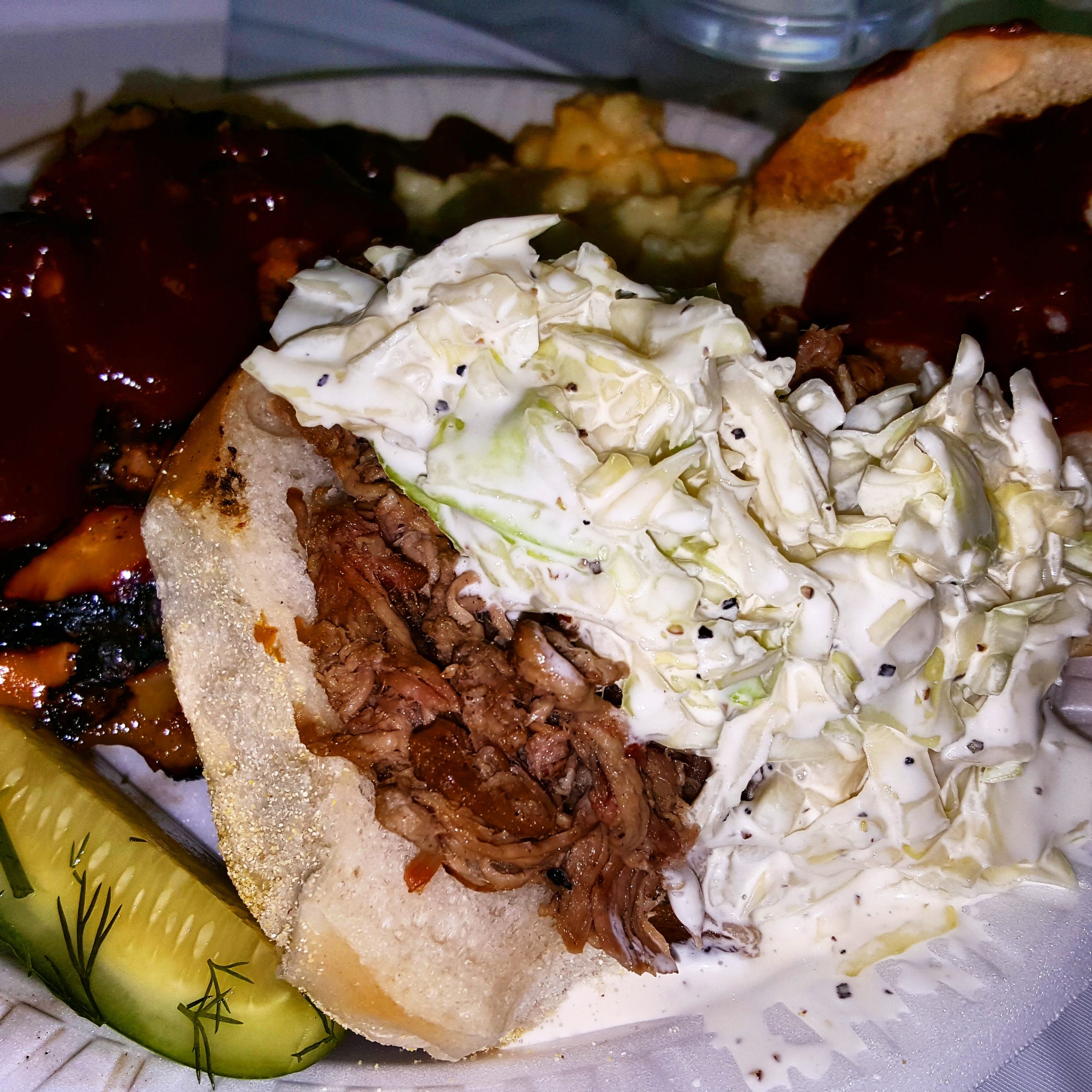 A dinner plate piled high with a pulled pork BBQ sandwich, baked beans, pickle, macaroni and cheese, beef brisket and barbecued chicken.